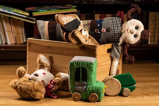 Children toys in a wooden box, books in the background on wooden floor. Teddy bear, wooden dog toy, rag doll, toy car, wooden green frog.